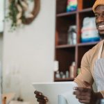 Barista with apron and beanie smiling as he takes an order from a customer in a cozy café setting.