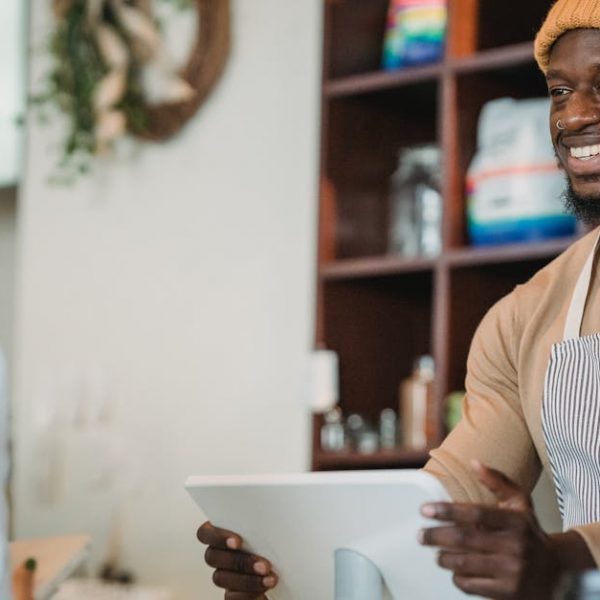 Barista with apron and beanie smiling as he takes an order from a customer in a cozy café setting.