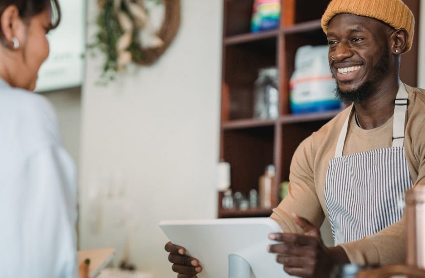 Barista with apron and beanie smiling as he takes an order from a customer in a cozy café setting.